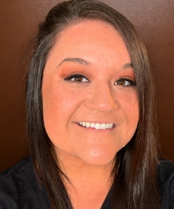 A woman with straight brown hair smiles at the camera. She is wearing a black top and has natural makeup with brown eyeshadow. The background is plain and brown.